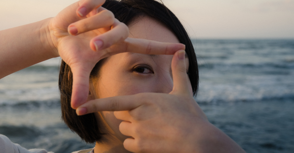 A close up of women standing by the ocean, looking at the camera through her index fingers and thumbs joined together to make a rectangle. 